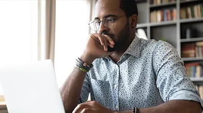 A man sitting in a library while using a laptop and resting his hand on his face 